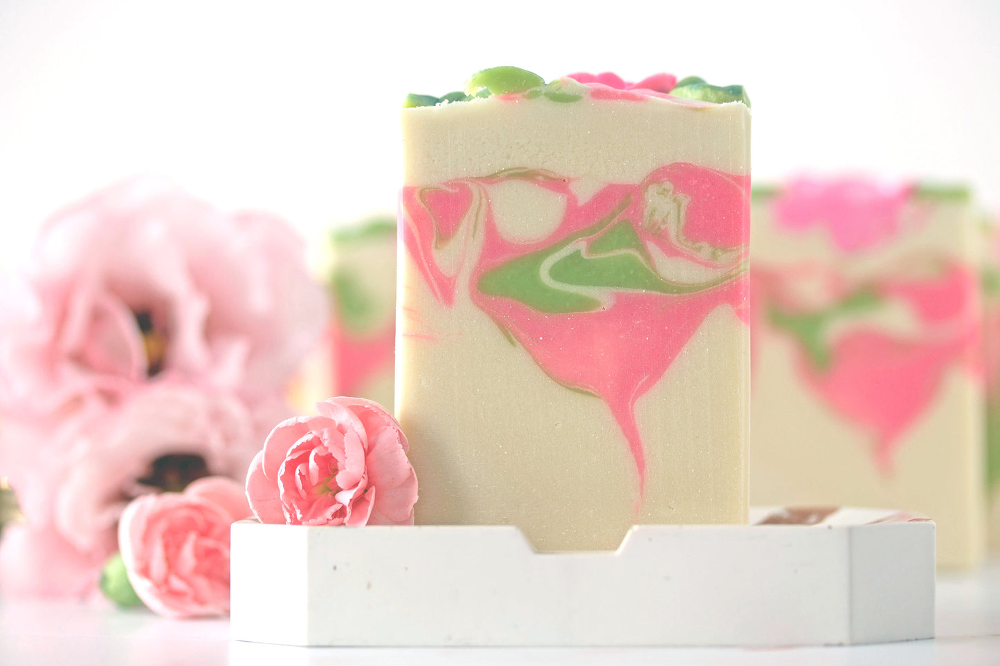 Bar of soap with pink and green swirls on a white stand, with pink flowers in the background.