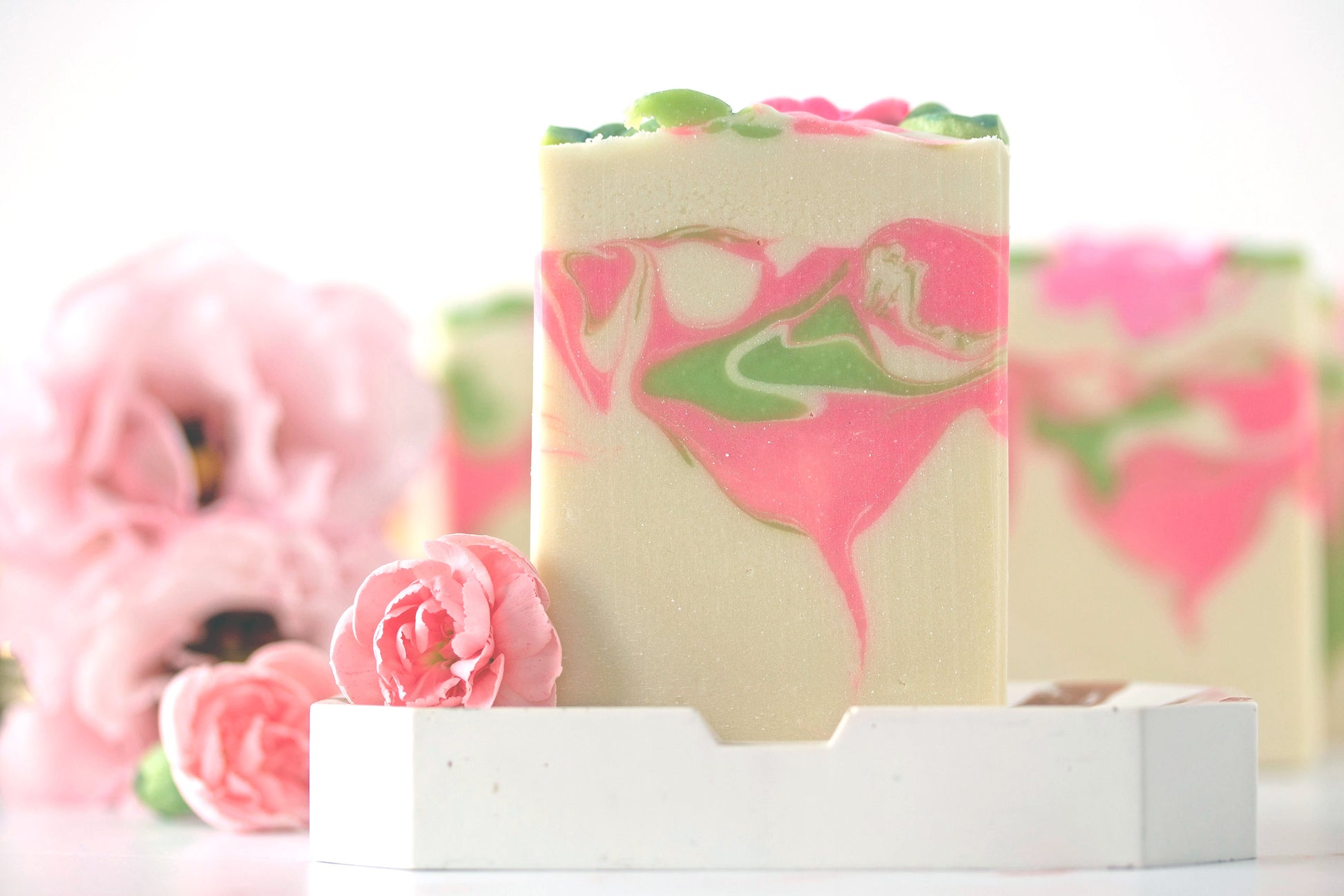 Bar of soap with pink and green swirls on a white stand, with pink flowers in the background.